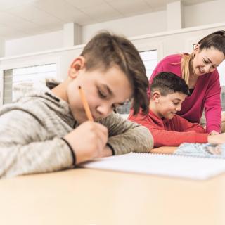 Lehrerin erklärt einem Schüler Unterrichtsstoff in einer Unterrichtssituation. Im Vordergrund sitzt ein anderer, schreibender Schüler.