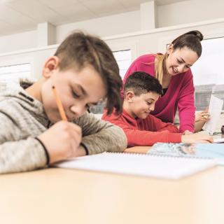 Lehrerin erklärt einem Schüler Unterrichtsstoff in einer Unterrichtssituation. Im Vordergrund sitzt ein anderer, schreibender Schüler.