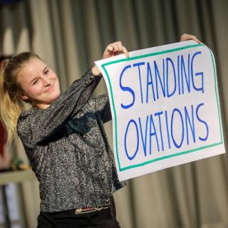 Jugendliche hält in handgeschriebenes Plakat mit dem Schriftzug "Standing Ovations" lächelnd hoch. 