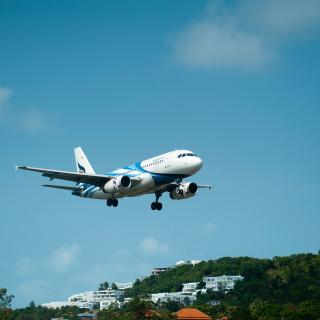 Blauer Himmel, davor startendes oder landendes Flugzeug, im Hintergrund bewaldete Landschaft mit weißen Häusern.