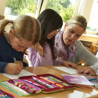 Eine Lehrerin sitzt zwischen Grundschülerinnen und erklärt etwas.