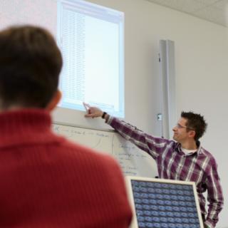 Unterrichtsituation mit digitalen Medien: Lehrer erklärt etwas an einem Smartboard, im Vordergrund steht ein Computerbildschirm.