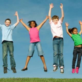 SiebenSieben Kinder unterschiedlichen Alters und verschiedener Herkunft springen in Jeans und T-Shirt vor sommerlich blauem Himmel auf einer gemähten Wiese mit erhobenden Armen in die Luft