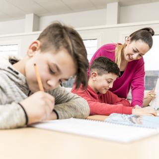 Unterrichtssituation: zwei schreibende Schüler, eine Lehrerin beugt sich über einen der beiden.