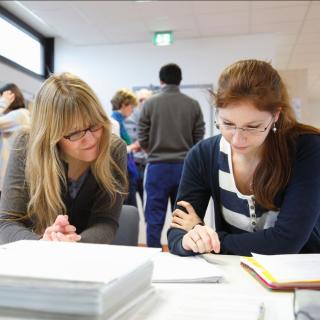 Zwei Lehrerinnen sitzen in einem Lehrerzimmer über Unterlagen gebeugt an einem Tisch.