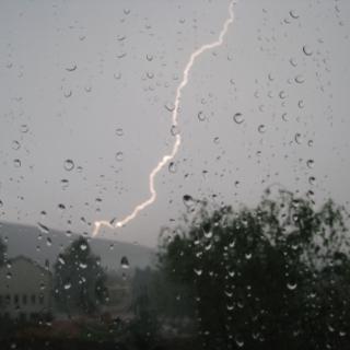 Sicht durch eine Fensterscheibe auf ein Unwetter mit Regen und Blitz, Bäume und ein Haus sind auszumachen.